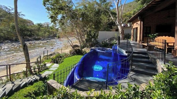 Un jacuzzi con tapa azul, rodeado por un sendero de piedra y vegetación, cerca de un edificio de madera con balcón.