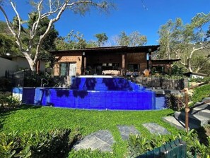 A swimming pool with blue tiles, surrounded by greenery and a wooden building with a tiled roof.