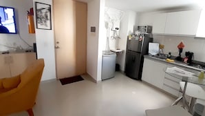 Een compacte keuken met een koelkast, magnetron en spoelbak. Een stoel is te zien op de voorgrond.