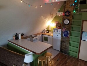 Compact kitchen with a small island counter, stove, sink, and refrigerator, featuring a green accent wall and string lights.