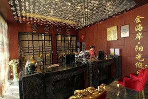 A reception desk with a person standing behind it, a red chair to the right, and traditional wall decorations. - Jinhai'an Sunshine Holiday Hotel (Yongfu)