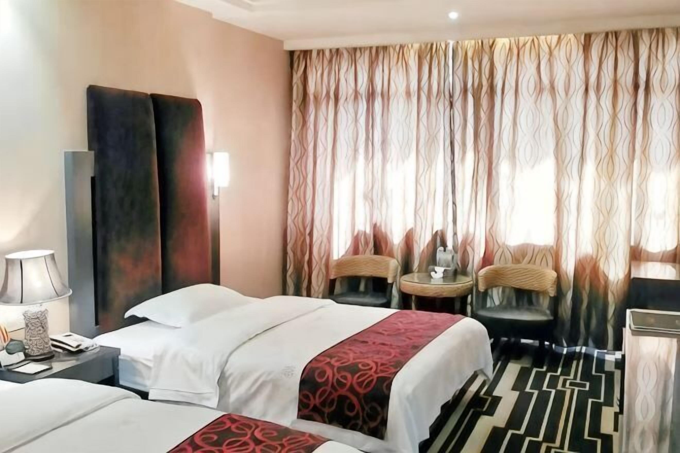 Ett hotellrum med två sängar, ett skrivbord, en stol, ett litet bord och ett stort fönster med gardiner.