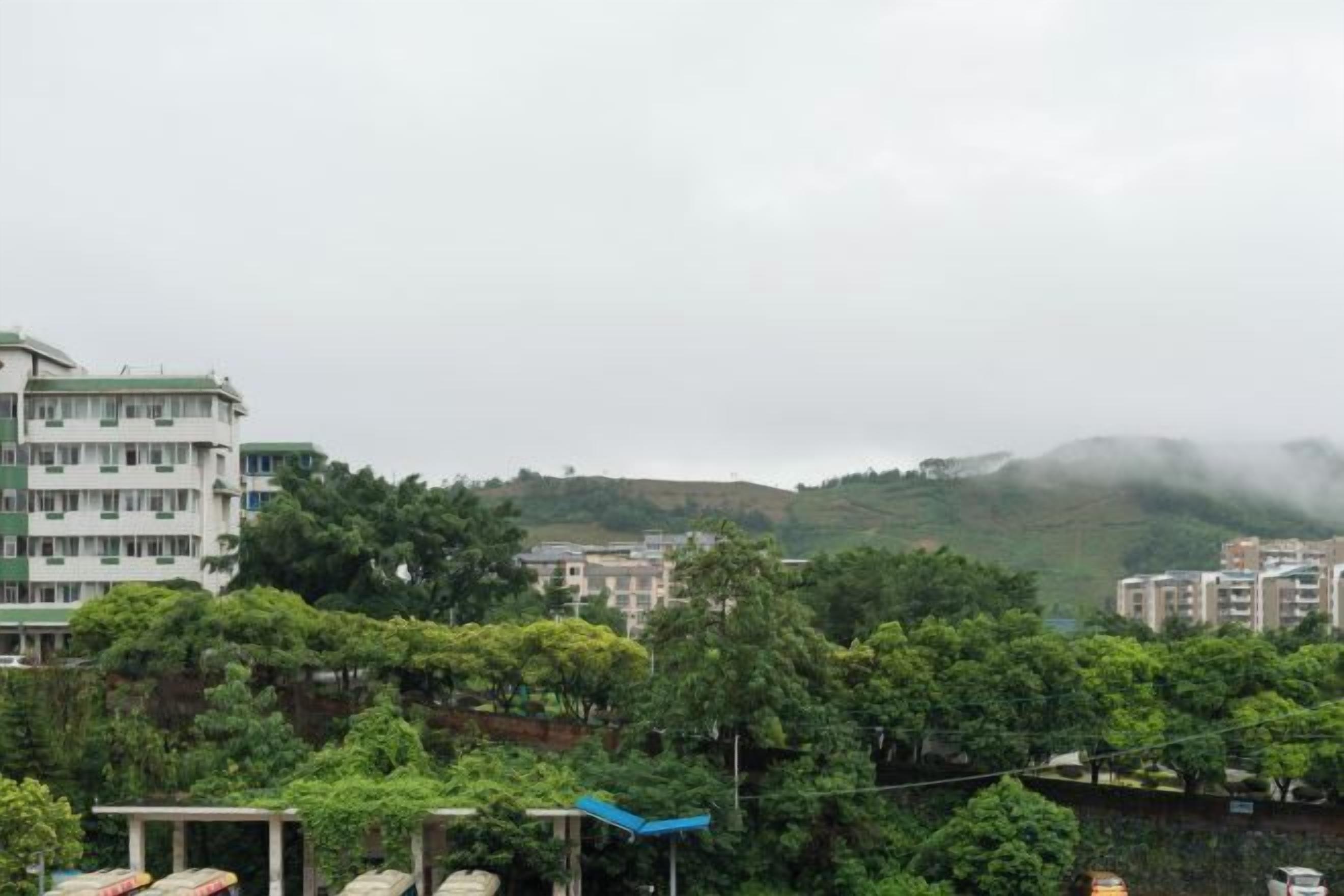 A multi-story building with a flat roof and a balcony, surrounded by trees and hills in the background.