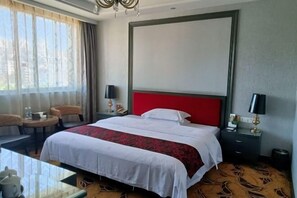 Habitación de hotel con una cama grande, cabecera roja, dos mesitas de noche con lámparas, una pequeña mesa redonda con una silla y vistas al exterior a través de amplias ventanas.