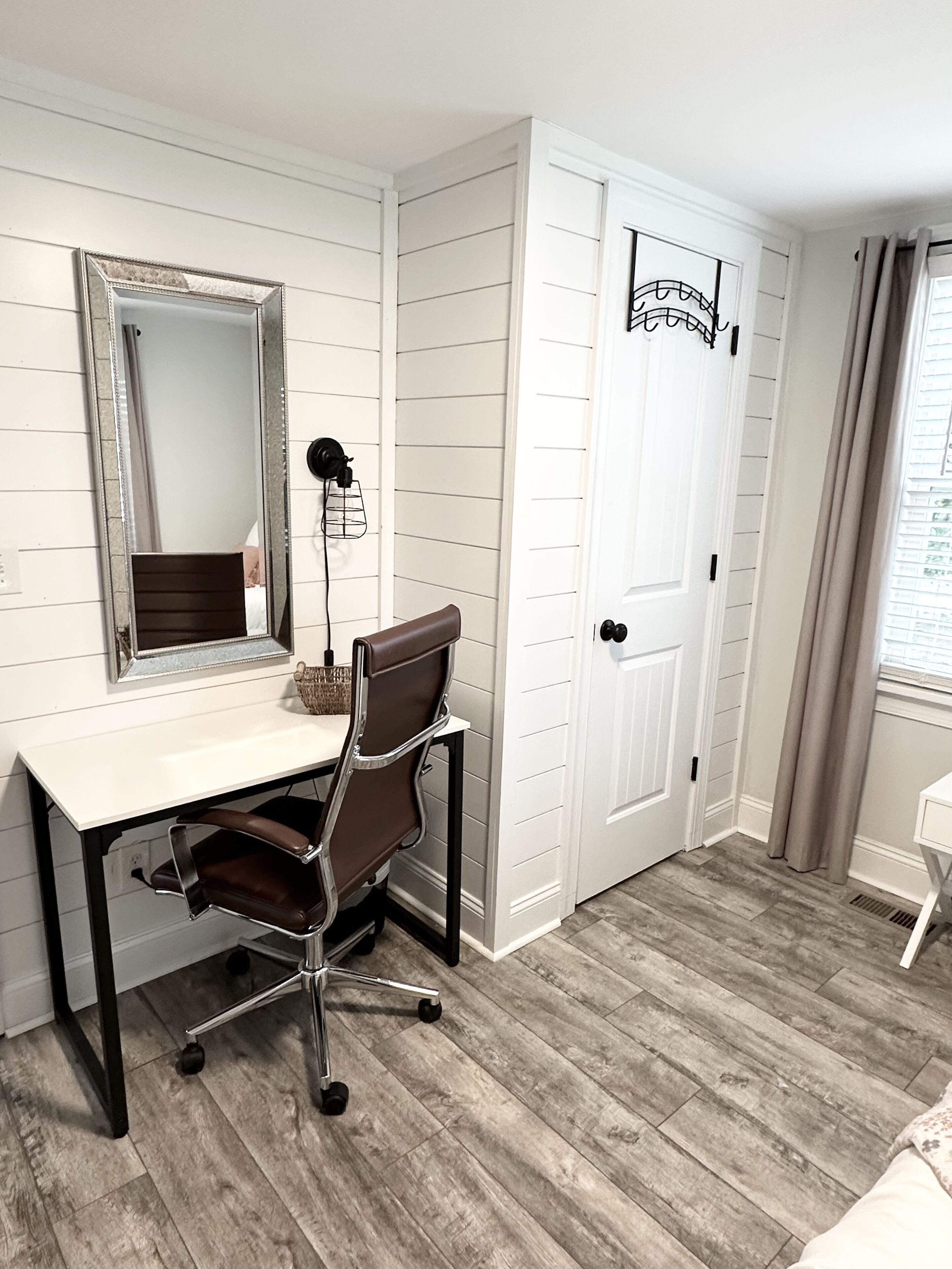 Una habitación con un escritorio blanco, una silla de oficina marrón, un espejo grande y una puerta con un letrero de "Welcome".