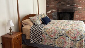 Une chambre avec un lit en bois, une couvre-lit à motifs, une petite table de chevet en bois avec une lampe, un mur en briques et une cheminée.