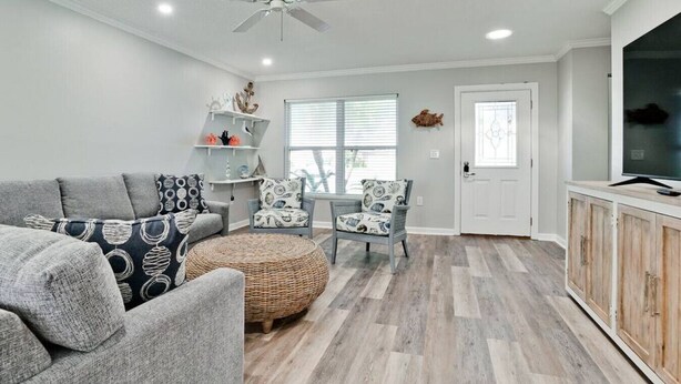 A living room with a grey sofa, patterned cushions, a wicker ottoman, a ceiling fan, and a TV on a wooden cabinet. - 19th - Aunies Place · Totally Updated Pool/FUN/Beaches (Bradenton)