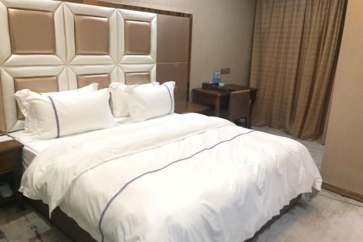 A hotel room with a large bed, a headboard with a geometric design, a nightstand, a small table, and a curtain.