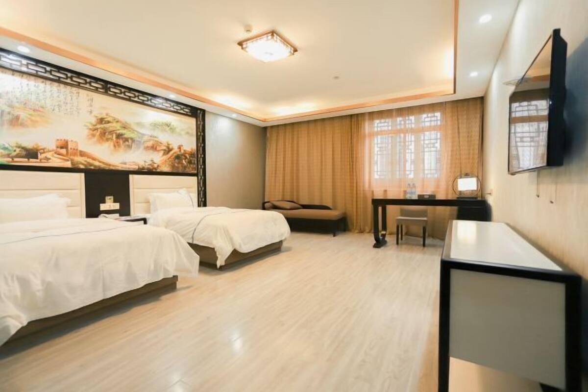 A hotel room with two beds, a desk, a chair, a TV, and a large painting on the wall.