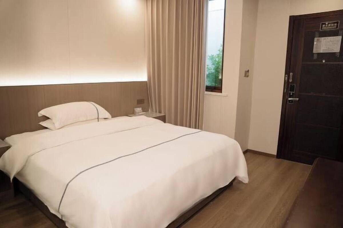 A hotel room with a bed, a nightstand, a window with curtains, and a door.