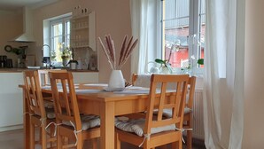 A dining area with a wooden table and chairs, a vase with dried plants, and a view of a red building through the window. - Vacation near Oldenburg and North Lake, garden, children, beach club, water skiing, golf (Rastede)