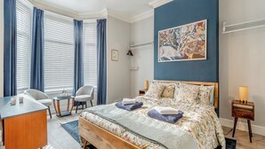 A bedroom with a bed, a desk, a chair, a window with curtains, a painting on the wall, and a bedside table with a lamp. - This ground floor apartment is less than a 10-minute walk away from the beach. (Scarborough)