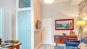 A living room with a blue sofa, a wooden dining table with plates, a television on the wall, and a painting above the sofa. - This ground floor apartment is less than a 10-minute walk away from the beach. (Scarborough)
