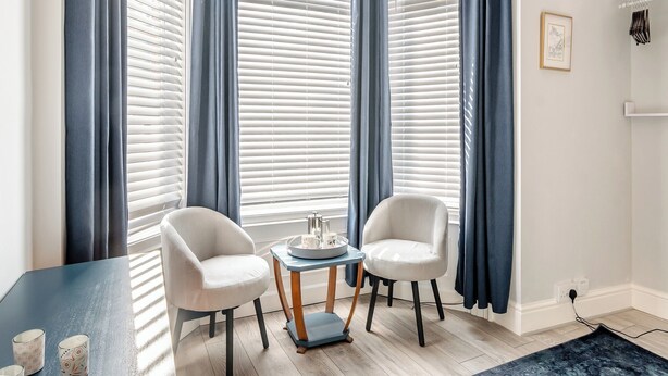 A room with two white chairs, a small table with a tray, and a window with blinds and blue curtains. - This ground floor apartment is less than a 10-minute walk away from the beach. (Scarborough)