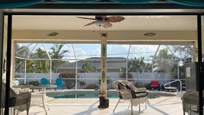 Una stanza con un'ampia porta scorrevole in vetro che si apre su una zona piscina, mobili in vimini e una ventola a soffitto.