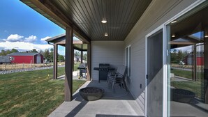 A covered patio with a grill and outdoor seating, a large planter, and a view of a red building in the background.