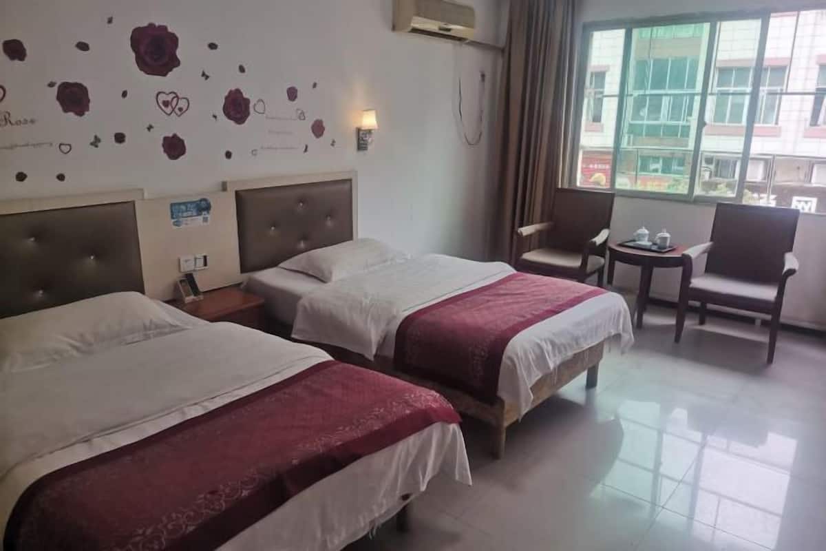 A hotel room with two beds, a small table with a cup and saucer, a chair, and a window with a view of buildings.