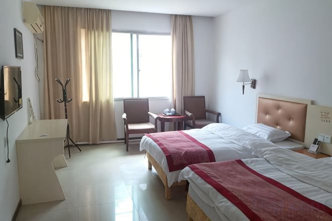 A hotel room with two beds, a desk, a chair, a window with curtains, and a lamp on the wall.