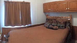 A bedroom with a single bed, brown bedding, a window with brown curtains, and wooden cabinets above the bed. - Gwin's Lodge Poplar Chalet #2 (Cooper Landing)