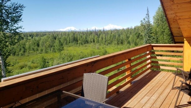 Une terrasse en bois avec une table et une chaise, offrant une vue sur un paysage boisé et des montagnes lointaines.