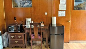 Il s’agit d’une pièce de taille modeste, équipée d’un bureau en bois, d’un four à micro-ondes, d’une machine à café, d’un réfrigérateur et d’une chaise. Deux tableaux encadrés ornent les murs.