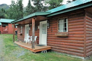 Une cabane rustique en rondins de bois, dotée d’un porche, de chaises en plastique blanc et d’une terrasse en bois.