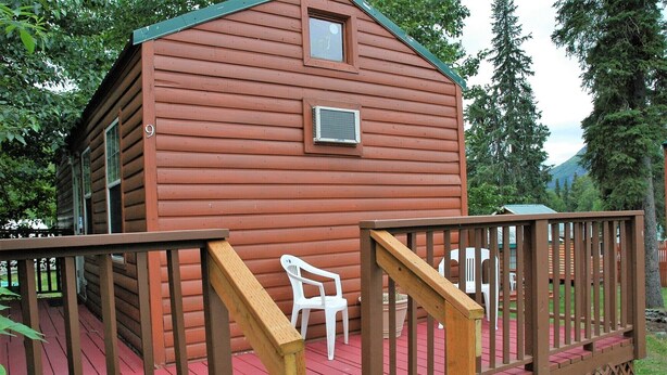 Una cabaña de madera con fachada roja, un pequeño porche con una silla blanca y una barandilla de madera.