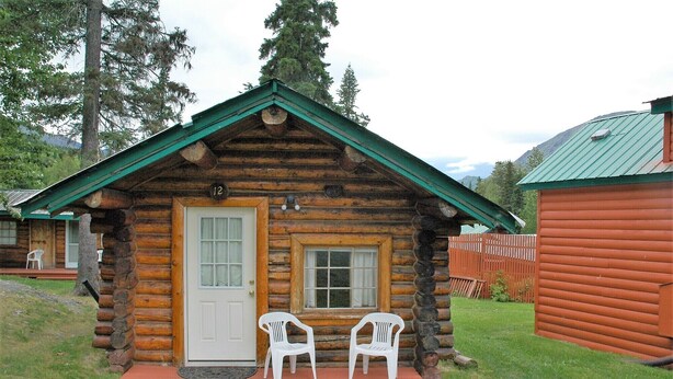 Une cabane en rondins avec un toit vert, une porte d’entrée et deux chaises en plastique blanches sur le porche.