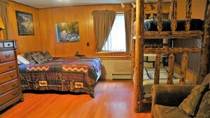 Ein gemütliches Zimmer mit Holzboden, einem Bett mit gemusterten Betttextilien, einem Schmuckkästchen, einem Fenster mit Vorhängen und einem rustikalen Sessel.