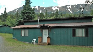 Eine grüne Holzhütte mit Veranda, einem weißen Plastikstuhl und Bergelandschaft im Hintergrund.