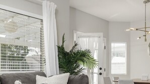Un salon moderne avec un canapé gris, des coussins blancs, une grande fenêtre avec des stores, un coin repas avec une table et des chaises, et une plante en pot.