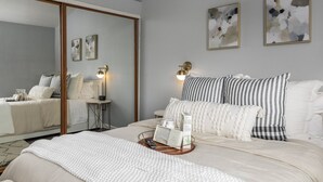 Une chambre avec un grand miroir, une table de chevet avec une lampe et deux tableaux abstraits accrochés au mur.