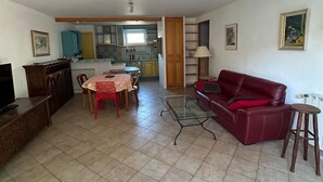 Una sala de estar con un sofá rojo, una mesa de centro de vidrio y una mesa de comedor con sillas. Se observa una cocina al fondo con armarios y una ventana.
