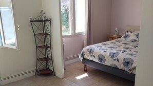Un dormitorio con cama, una mesita de noche con lámpara, una ventana con cortinas y una estantería metálica.