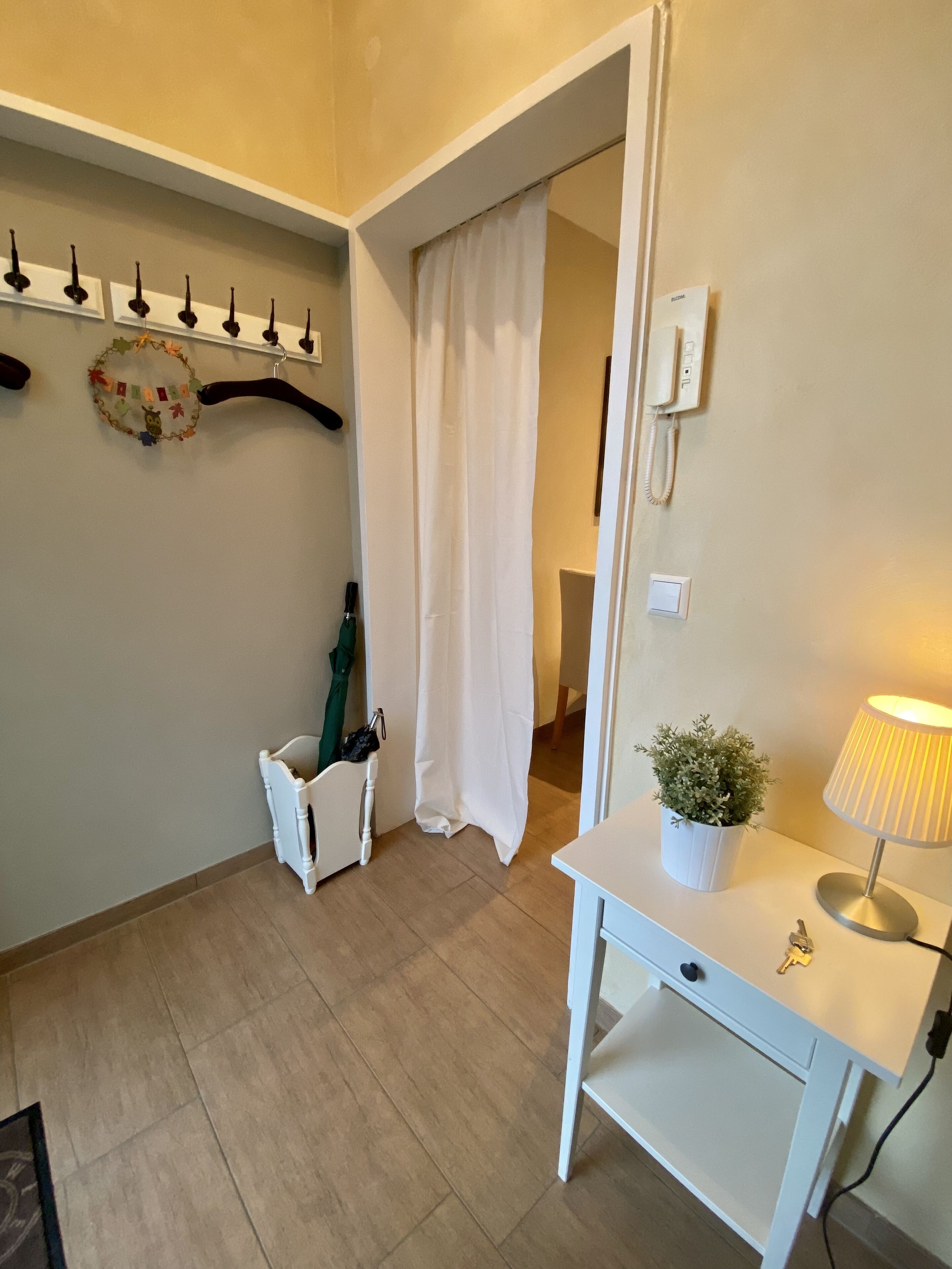 Ett hotellrum med ett vitt skrivbord, en lampa, en krukväxt, en klädställning med galgar och en dörr med en vit gardin.