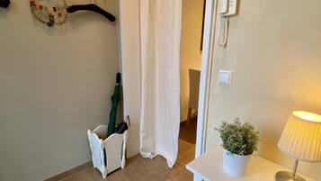 Ett hotellrum med ett vitt skrivbord, en lampa, en krukväxt, en klädställning med galgar och en dörr med en vit gardin.