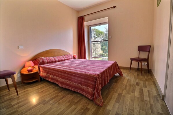 Un dormitorio con una cama de madera, una mesita de noche con lámpara, una silla y una ventana con vista a árboles.