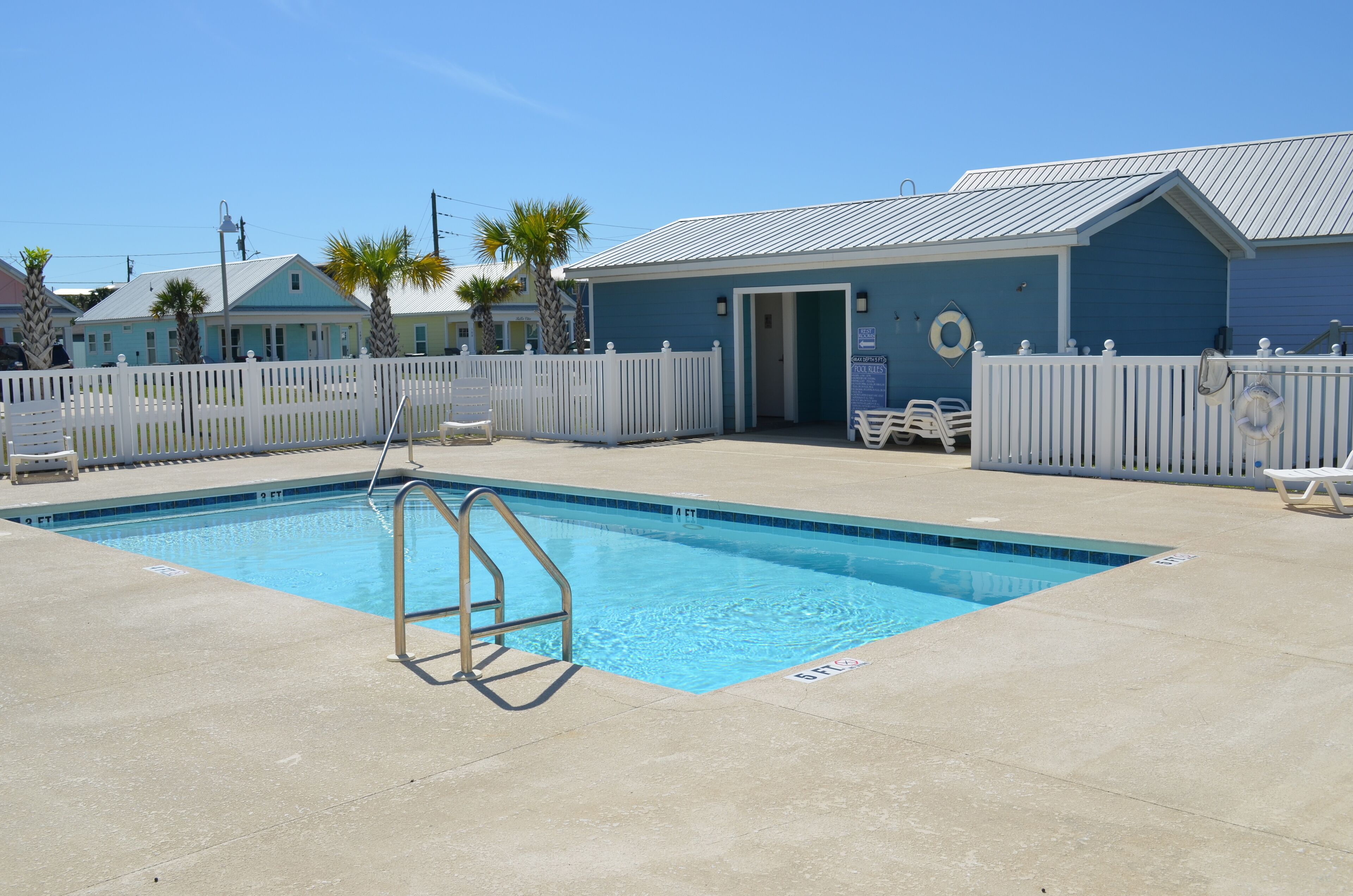 A swimming pool with a white fence, a lifeguard stand, and a blue building with a door.