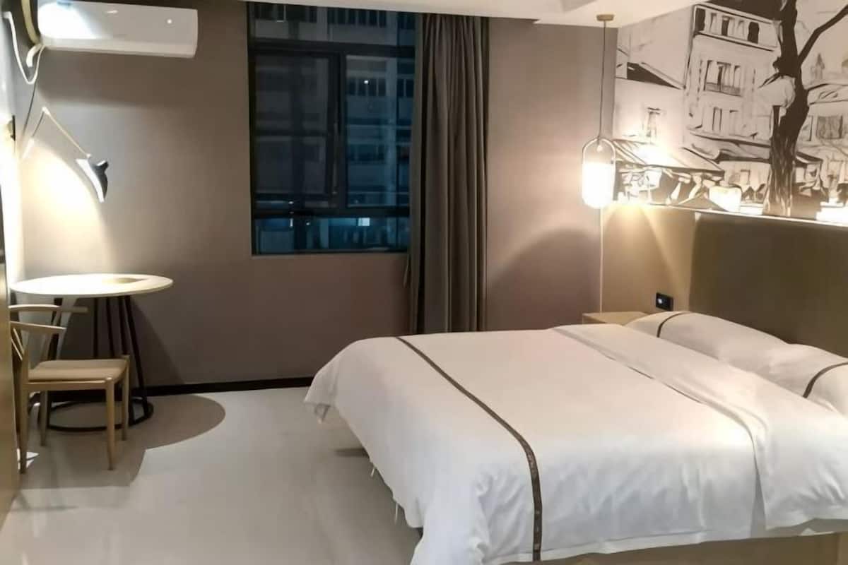 A hotel room with a bed, a round table with chairs, a window, and a wall-mounted lamp.