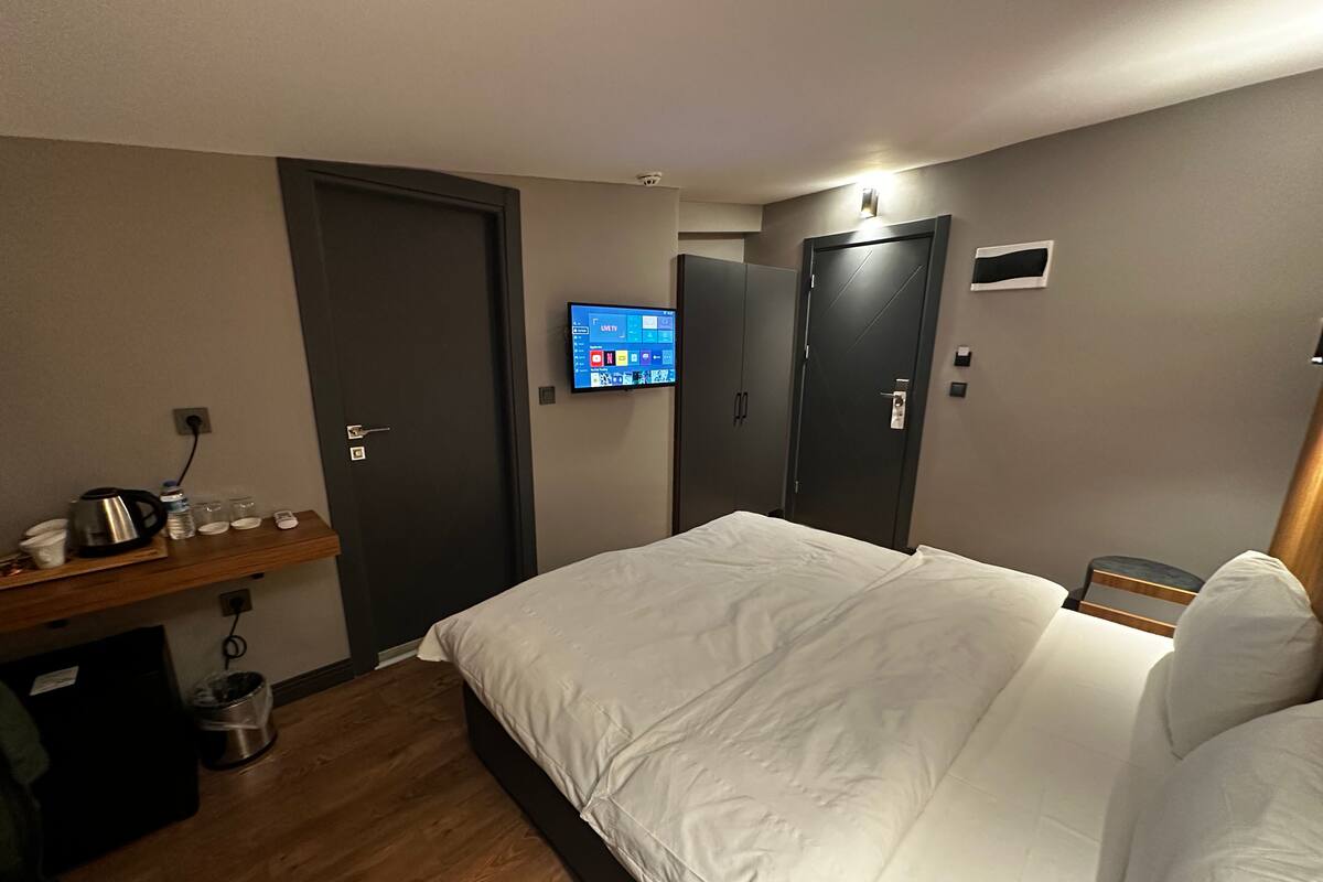 A hotel room with a bed, a TV mounted on the wall, a wooden shelf with toiletries, and a door.