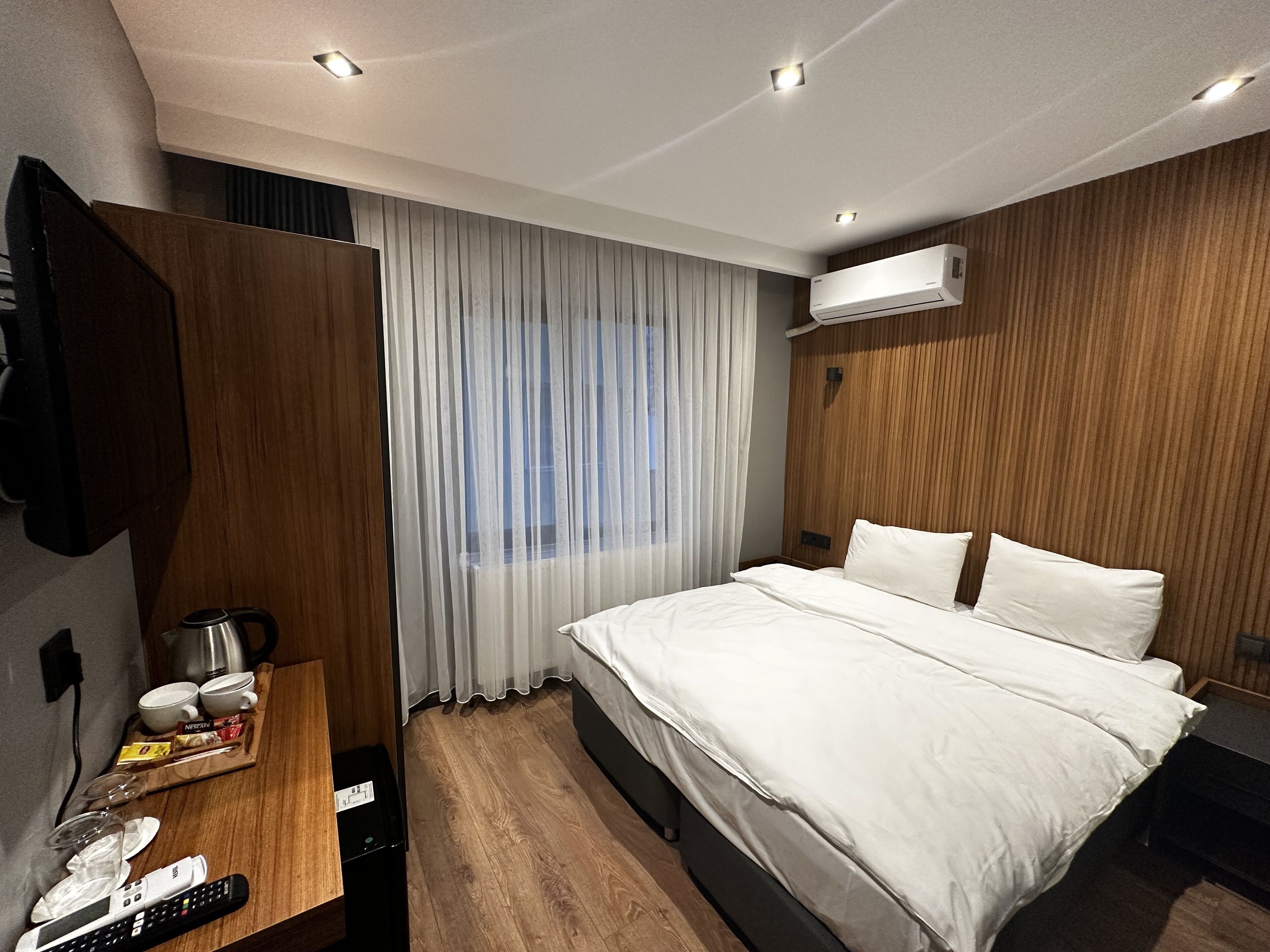 A modern hotel room with a large bed, wooden paneling, a desk with a kettle, cups, and a remote control.