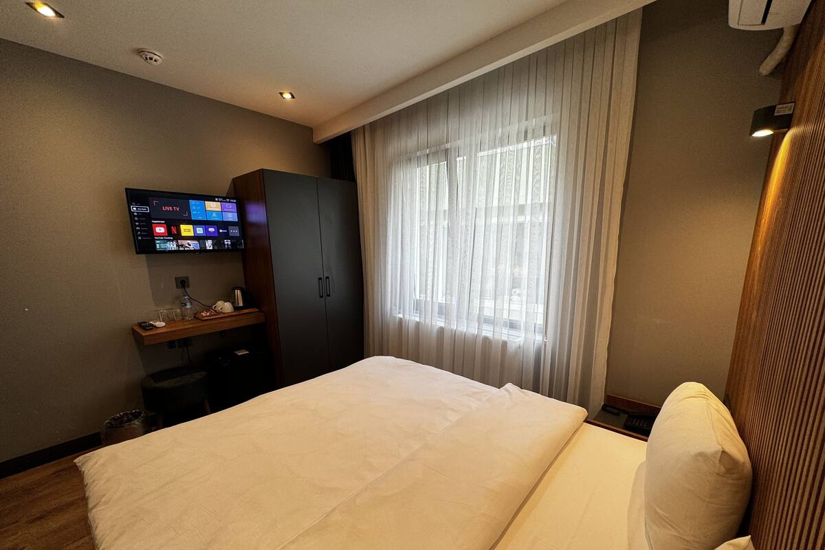 A hotel room with a large bed, a TV mounted on the wall, a wardrobe, and a window with curtains.
