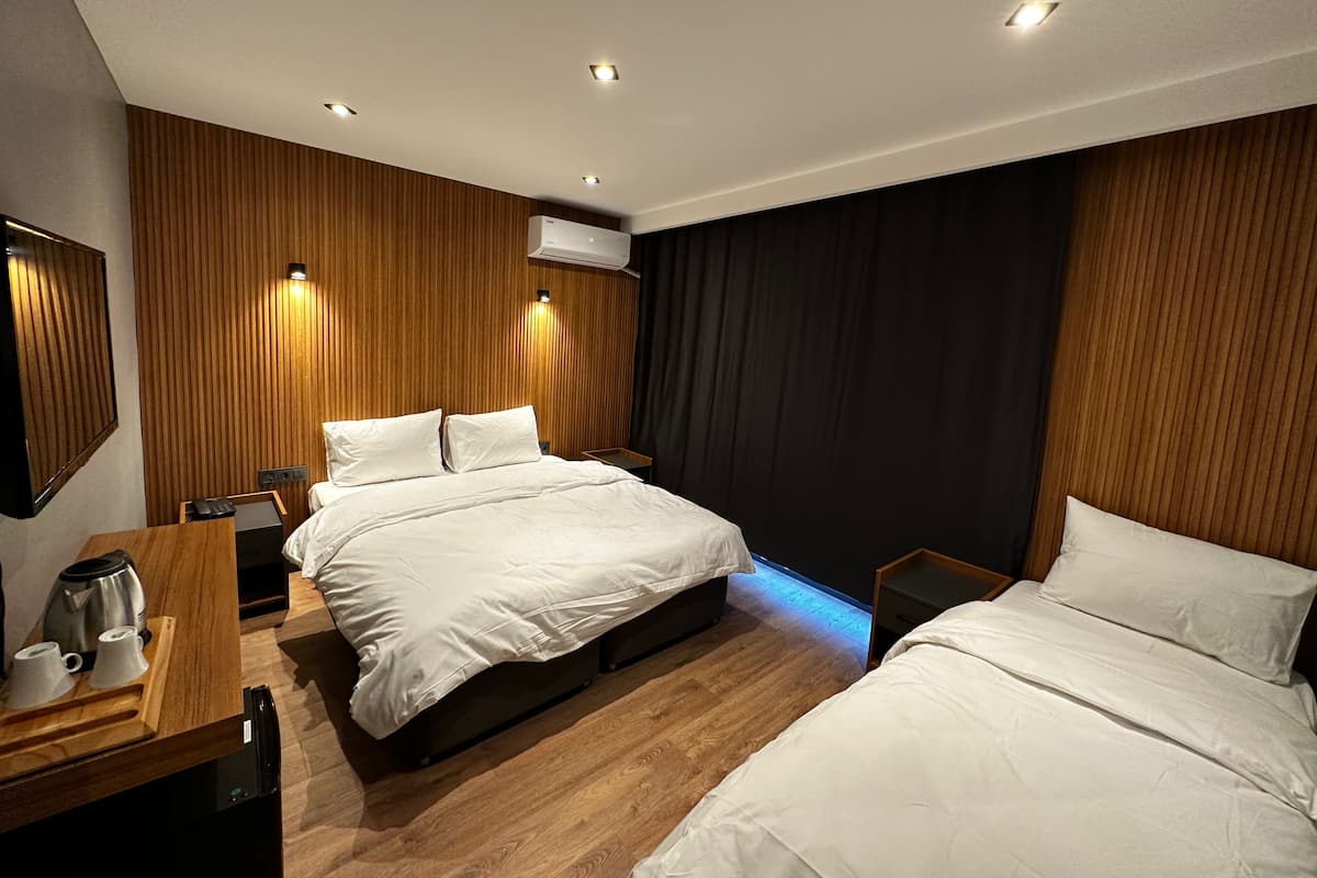 A modern hotel room with two beds, wooden panel walls, recessed ceiling lights, an air conditioning unit, a bedside table with a coffee maker, and a small desk with a telephone.