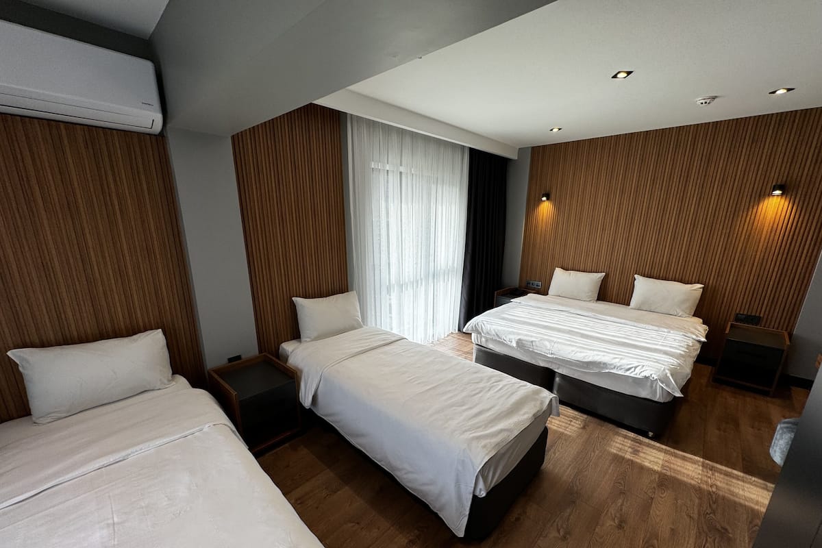 A modern hotel room with two beds, wooden paneling, a large window with curtains, and built-in lighting.
