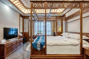 Un lit traditionnel à baldaquin, avec un téléviseur posé sur une console en bois, un bureau avec une chaise et un paravent décoratif.