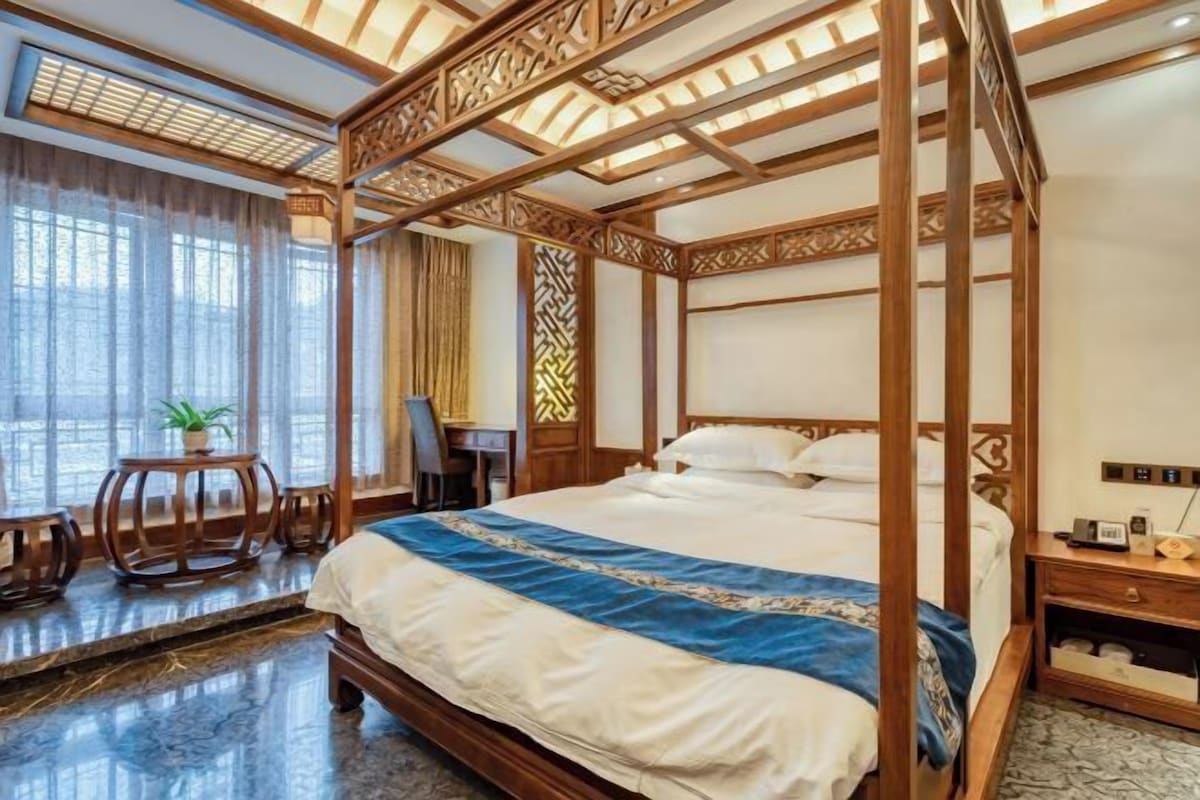 Un lit à baldaquin avec une couvre-lit bleue et blanche, un bureau en bois avec une chaise, une plante en pot et une grande fenêtre avec des rideaux légers.