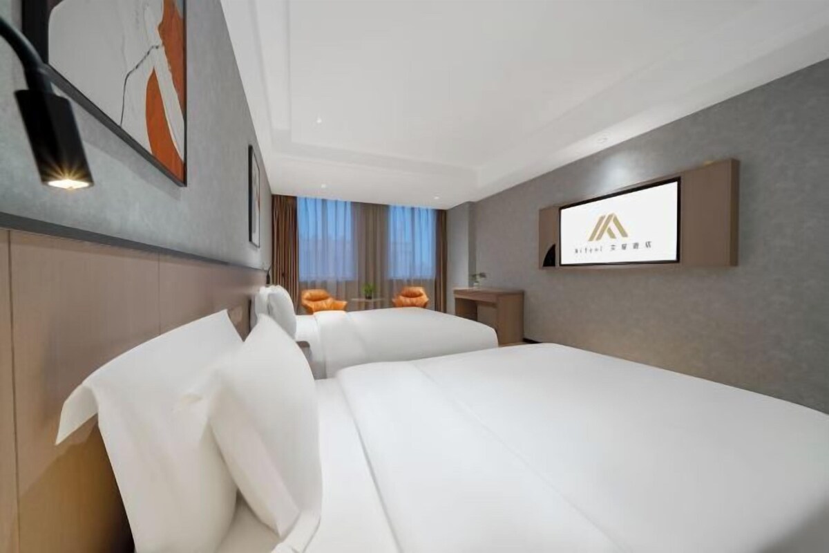 Ein modernes Hotelzimmer mit einem großen Bett, einem Schreibtisch und einem Sessel. Ein Fernseher ist an der Wand befestigt und es gibt ein Fenster mit Vorhängen.