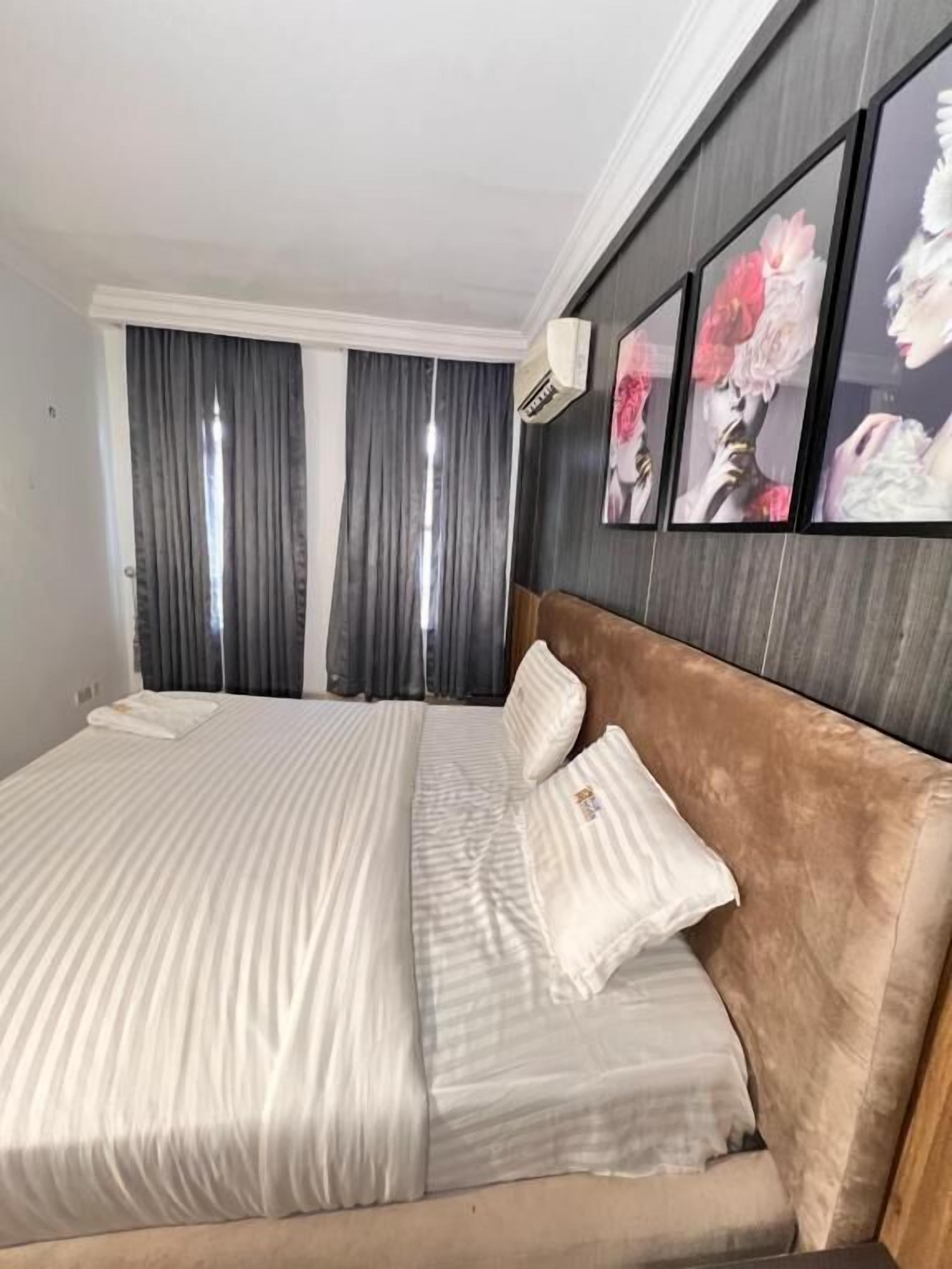 A neatly made bed with striped bedding and a brown headboard, a large window with grey curtains, and three framed floral artworks on the wall.