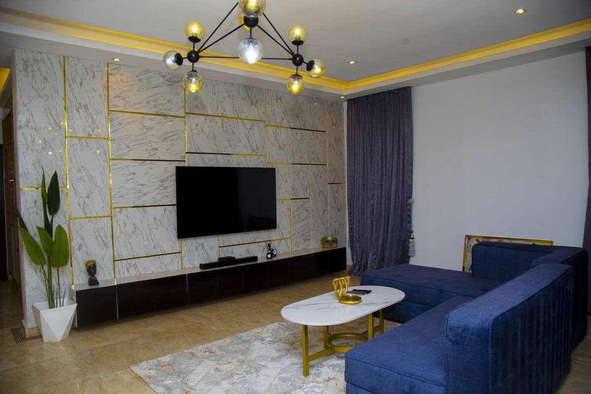 Una sala de estar moderna con un televisor de pantalla plana grande, un sofá azul, una mesa de centro blanca y una planta decorativa.