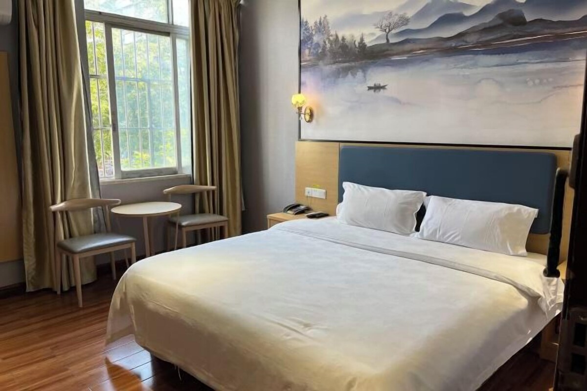 Une chambre d’hôtel avec un grand lit, une petite table, deux chaises, une table de chevet et un tableau représentant un paysage.
